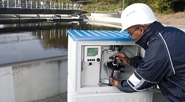 A person in a white helmet and dark jacket operating equipment housed within a small white and blue structure, located beside a water body with industrial elements in the background. The equipment is housed within a small white structure with a blue roof, which appears to be some sort of control or monitoring station.
