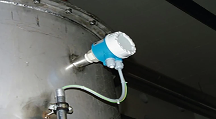 A cylindrical blue sensor is attached to a large metallic tank with rivets. The sensor is connected to wires and the background is dark. The image appears to be taken in an industrial or manufacturing facility.