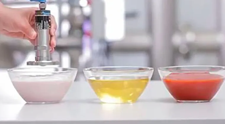 A person’s hands are using a hand blender to mix a white creamy substance in a clear glass bowl. Two other clear glass bowls containing yellow and red liquids are on the countertop. The background is blurred but shows part of a clean, modern kitchen with stainless steel appliances.