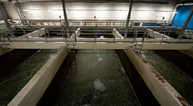 An indoor wastewater treatment facility with large open tanks containing greenish water and walkways above.