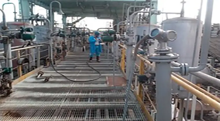 A worker in blue uniform inspecting equipment at an industrial plant with various pipes and machinery.